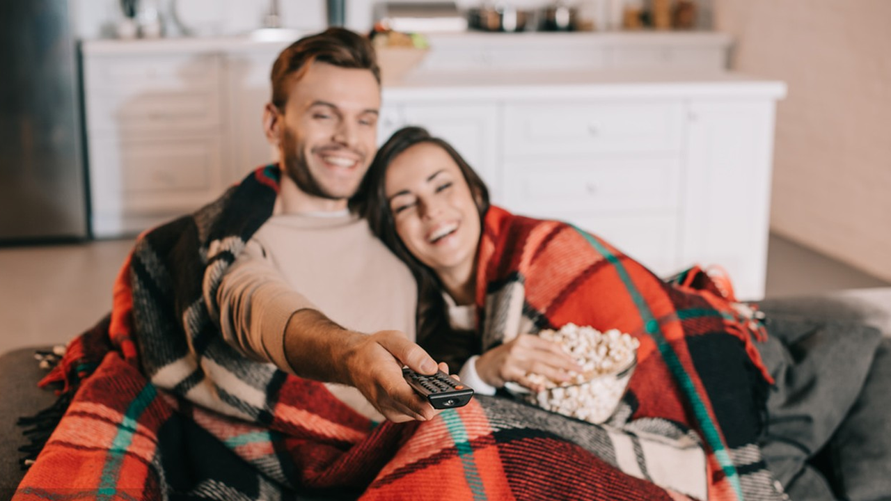 A cozy scene showing two people relaxing under a red plaid blanket while watching something, presumably TV or a movie, with a bowl of popcorn.