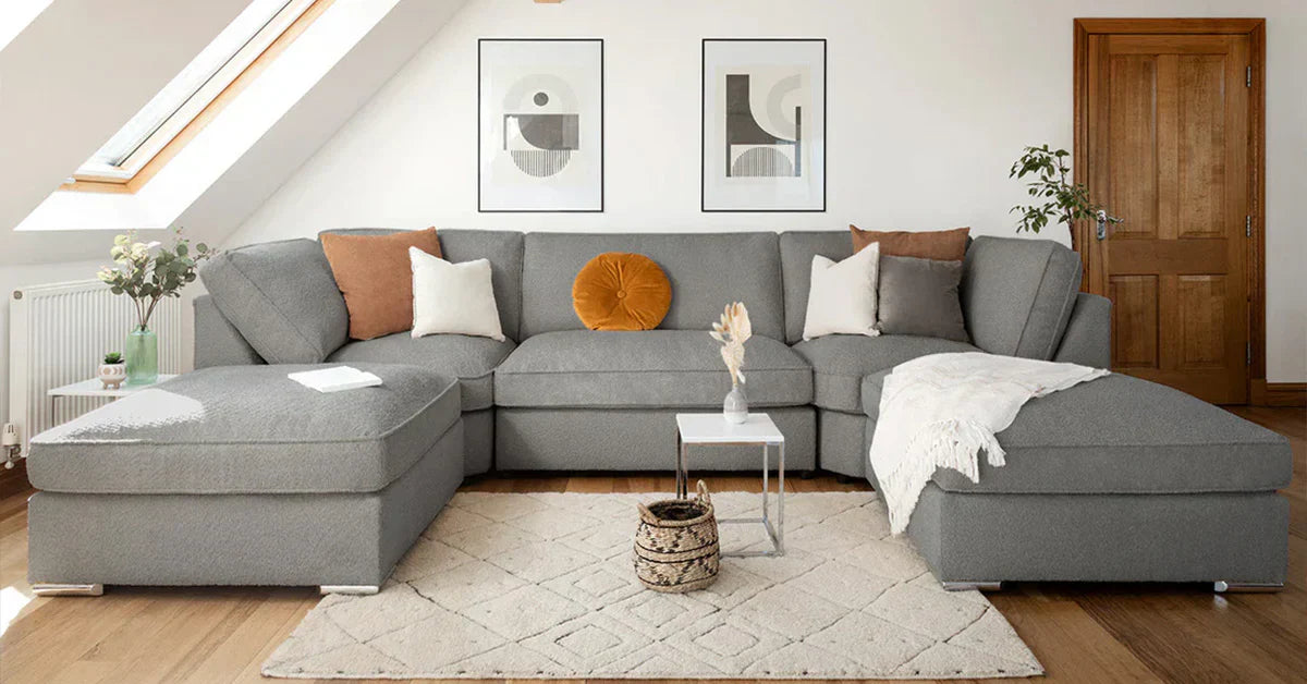Modern gray L-shaped sectional sofa with hidden storage ottoman, caramel and mustard throw pillows, white walls with geometric art, natural light from skylight, wooden floors with neutral rug, and minimalist decor.
